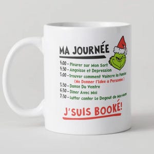 Peut inclure: Tasse en céramique blanche avec un design humoristique. La tasse présente le texte "MA JOURNÉE" et un emploi du temps d'activités, dont "Pleurer sur mon sort" et "Danser avec moi". Un personnage de dessin animé vert avec un chapeau de Père Noël est également sur la tasse.