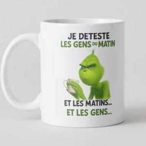 Peut inclure: Mug en céramique blanche avec un personnage de dessin animé vert grincheux tenant une tasse. Le texte "JE DETESTE LES GENS DU MATIN ET LES MATINS... ET LES GENS..." est écrit en vert, une blague sur les matins.