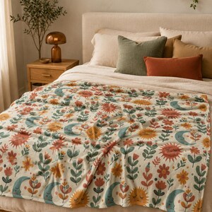 May include: A cream-colored throw blanket featuring a repeating pattern of stylized suns with faces, crescent moons with faces, and various floral elements in shades of orange, red, teal, and green, draped over a bed with neutral-toned pillows.