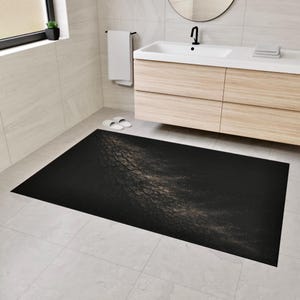 May include: A rectangular black bath mat with a textured, tree bark-like pattern in shades of brown and gold. The mat is placed on a tiled floor in a bathroom setting, near a sink and cabinet.