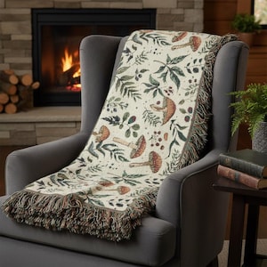 Puede incluir: Una manta crema sobre un sillón gris. La manta presenta un patrón repetido de setas, hojas y bayas en tonos marrones, verdes y rojos. La manta tiene un borde con flecos.
