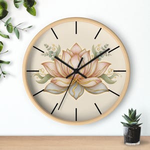 Pode incluir: Um relógio de parede redondo com moldura de madeira clara. O mostrador do relógio apresenta uma ilustração detalhada de uma flor de lótus em tons de rosa, dourado e azul-acinzentado, com folhas verdes. Linhas pretas marcam as horas.