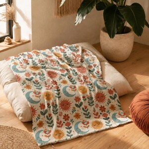 May include: A cream-colored throw blanket featuring a repeating pattern of smiling suns, crescent moons with faces, and various stylized flowers in shades of orange, yellow, teal, and muted red, printed across the soft fabric surface.