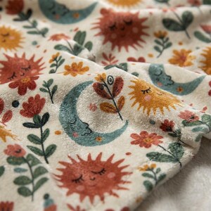 May include: A soft, cream-colored fabric blanket featuring a repeating pattern of smiling suns in shades of orange and yellow, teal crescent moons with faces, and various small flowers with green stems and leaves printed across the surface.