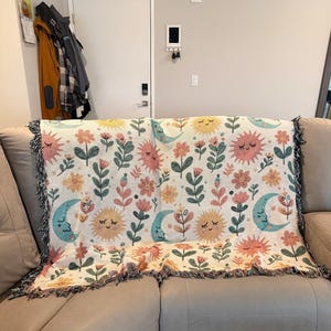 Puede incluir: Una manta crema con un patrón repetido de soles, lunas y flores sonrientes en colores pastel. La manta tiene un borde con flecos y está sobre un sofá.