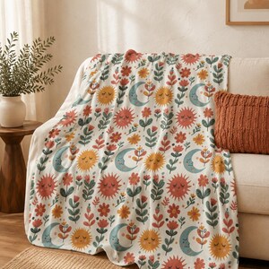 May include: A white throw blanket featuring a repeating pattern of smiling suns in shades of orange and yellow, crescent moons in light blue, and various stylized flowers in red, orange, and green. The fabric has a soft, plush texture throughout.
