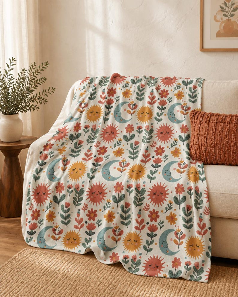 May include: A white throw blanket featuring a repeating pattern of smiling suns in shades of orange and yellow, crescent moons in light blue, and various stylized flowers in red, orange, and green. The fabric has a soft, plush texture throughout.