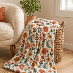 May include: A soft throw blanket featuring a repeating pattern of smiling suns, crescent moons, and various flowers in shades of terracotta, muted green, and soft blue on an off-white background, draped over a woven wicker basket.
