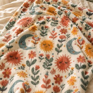 May include: A cream-colored fleece blanket featuring a repeating pattern of smiling suns in orange and yellow, crescent moons in muted blue, and various stylized flowers in shades of red, orange, and green, all with a soft, textured surface.