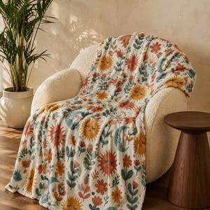 May include: A soft throw blanket featuring a repeating pattern of smiling suns, crescent moons, and various flowers in shades of orange, yellow, teal, and muted red on a cream-colored background, draped over a textured off-white armchair.