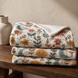 May include: A folded plush throw blanket featuring a repeating pattern of smiling suns, crescent moons, and various flowers in muted tones of orange, yellow, teal, and green on an off-white background with a soft white underside.