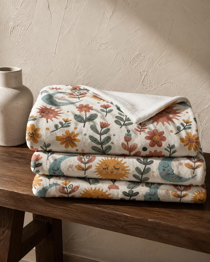 May include: A folded plush throw blanket featuring a repeating pattern of smiling suns, crescent moons, and various flowers in muted tones of orange, yellow, teal, and green on an off-white background with a soft white underside.