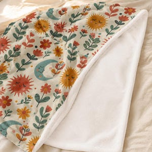 May include: A soft fleece blanket featuring a cream-colored background with a repeating pattern of smiling suns, crescent moons, and various red, orange, and green flowers. The underside of the blanket is a solid, plain white plush fabric.