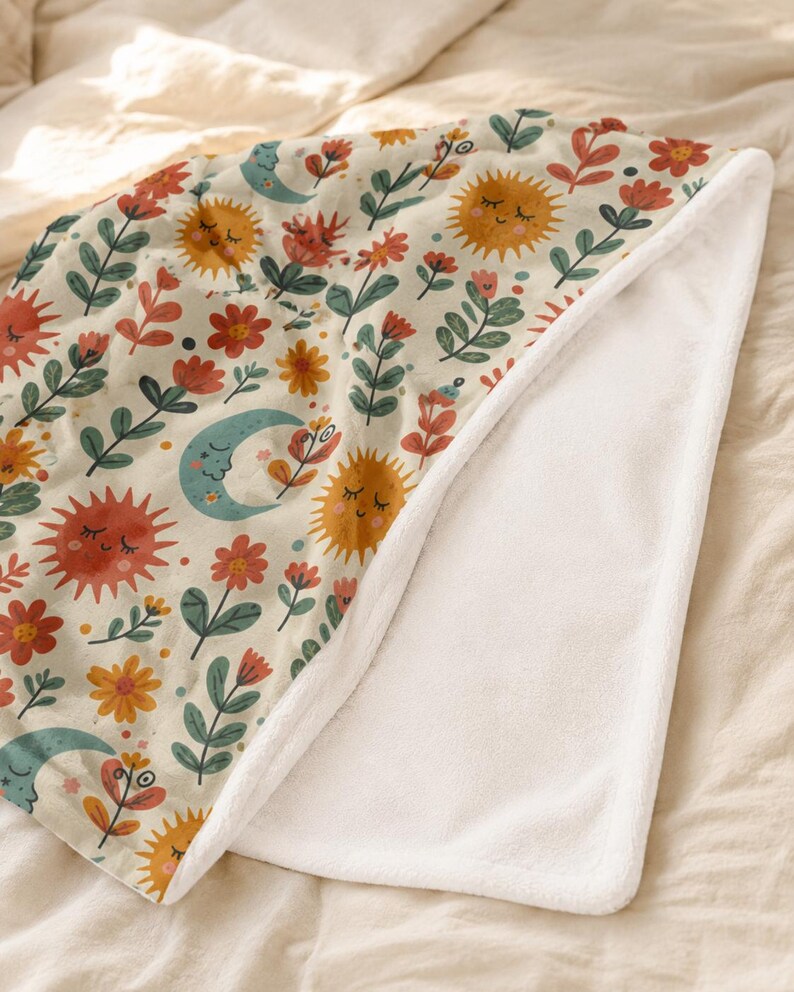 May include: A soft fleece blanket featuring a cream-colored background with a repeating pattern of smiling suns, crescent moons, and various red, orange, and green flowers. The underside of the blanket is a solid, plain white plush fabric.