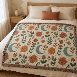 May include: A woven throw blanket featuring a repeating pattern of orange suns with faces, blue crescent moons, and various orange and green flowers on a cream background. The blanket has a fringed edge with multi-colored threads.