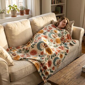 May include: A cream-colored throw blanket featuring a repeating pattern of orange suns, blue crescent moons with faces, and various red and orange flowers with green leaves, draped over a person resting on a beige upholstered sofa.