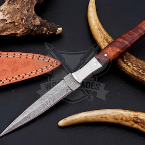 May include: A Damascus steel dagger with a wooden handle and a leather sheath. The blade has a detailed pattern, and the handle is a rich brown color. The sheath is a lighter brown with decorative stitching. The dagger is displayed with antlers.