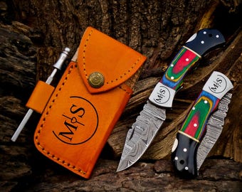 Puede incluir: Una funda de cuero naranja para cuchillo con un cierre de metal y las iniciales "M&S" está junto a dos navajas. Las navajas tienen hojas estampadas y mangos de madera coloridos. Una navaja está abierta y la otra cerrada.