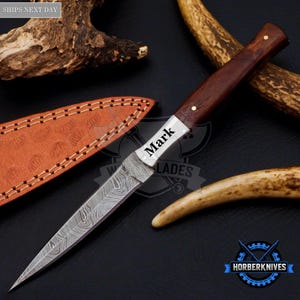May include: A fixed-blade knife with a Damascus steel blade featuring a wavy pattern and the name Mark engraved on the bolster. The handle is made of dark brown wood with a brass pin. It is accompanied by a tan leather sheath with stitching.