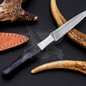 May include: A Damascus steel knife with a black handle and a leather sheath. The blade has a unique pattern. The knife is displayed with a piece of antler and a dark background. The text "WOOT BLADES" is visible.