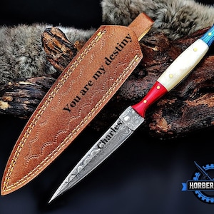 May include: A fixed-blade knife with a Damascus steel blade engraved with the name Charles, featuring a handle with red, white, and blue sections and brass pins. It includes a tan leather sheath embossed with the text You are my destiny.