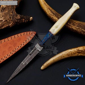 May include: A fixed-blade knife featuring a Damascus steel blade with the name Kevin engraved on it, a blue metal bolster, and a smooth cream-coloured handle. The knife is accompanied by a textured tan leather sheath with visible stitching.