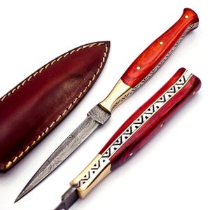 May include: A handcrafted dagger with a Damascus steel blade and a red wooden handle. The handle is accented with brass and decorative silver inlays. A brown leather sheath is included.