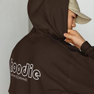Op de afbeelding: Een donkerbruine hoodie met het woord "hoodie" in het wit op de rug gedrukt. De tekst gaat vergezeld van een definitie. De hoodie heeft een capuchon en lange mouwen.