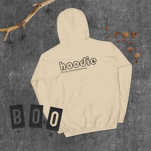 Op de afbeelding: Een crèmekleurige hoodie met het woord "hoodie" in het zwart gedrukt, samen met de uitspraak en definitie. Het woord "BOO" is in zwarte letters links onderaan gespeld. De achtergrond is donkergrijs.