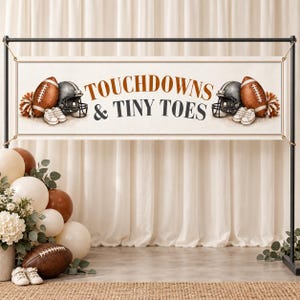 Op de afbeelding: Een rechthoekige banner met de tekst TOUCHDOWNS & TINY TOES in bruin en donkergrijs. Het ontwerp bevat illustraties van twee bruine footballs, twee donkergrijze footballhelmen, twee paar witte babyschoentjes en oranje pompons.