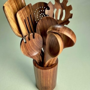 May include: A set of dark brown wooden kitchen utensils including a solid turner, a slotted turner, a pasta server, a skimmer, and two spoons, all housed in a matching cylindrical wooden utensil holder with a small circular logo on the front.