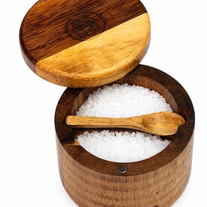 May include: A wooden salt cellar with a lid, filled with coarse white salt. The lid is two-toned, with a dark brown and light brown wood grain. A small wooden spoon rests inside. The words "Coki Home" are printed on the side.