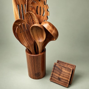 May include: A set of dark brown wooden kitchen utensils including a slotted turner, pasta server, slotted spoon, solid spoon, and ladle, all stored in a cylindrical wooden holder, accompanied by two square wooden coasters with a small circular logo.