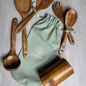 May include: A wooden kitchen utensil set includes a spatula, slotted spatula, fork, spoon, and ladle. The set is accompanied by a light green drawstring bag with the "Coki Home" logo and a wooden utensil holder. The utensils are made of a light brown wood.