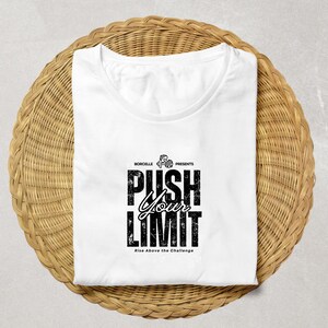 Puede incluir: Camiseta blanca con el texto "PUSH YOUR LIMIT" en una fuente negra desgastada. La camiseta tambi&eacute;n incluye el texto "Rise Above the Challenge" y la marca "BORCELLE PRESENTS". La camiseta est&aacute; doblada sobre una superficie circular tejida.