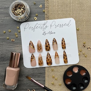 May include: A set of ten almond-shaped, press-on nails with a leopard print design, displayed on a white card. The card reads "Perfectly Pressed By Alena." Also shown are a bottle of nude nail polish, a makeup brush, a palette of nail colors, and a jar of gold glitter.