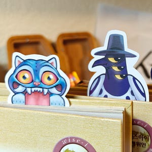 Puede incluir: Dos marcapáginas de personajes caprichosos. Uno es una criatura azul con forma de gato con grandes ojos anaranjados y dientes afilados. El otro es una figura púrpura que lleva un sombrero y un atuendo de lunares blancos y negros. Los marcapáginas están colocados en un libro.