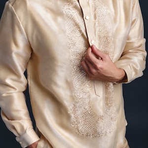 May include: A long-sleeved, champagne-colored barong tagalog shirt featuring a mandarin collar and a central vertical panel with intricate, tonal embroidery. The garment has a button-down front placket and cuffed sleeves, crafted from a semi-sheer, lustrous fabric.