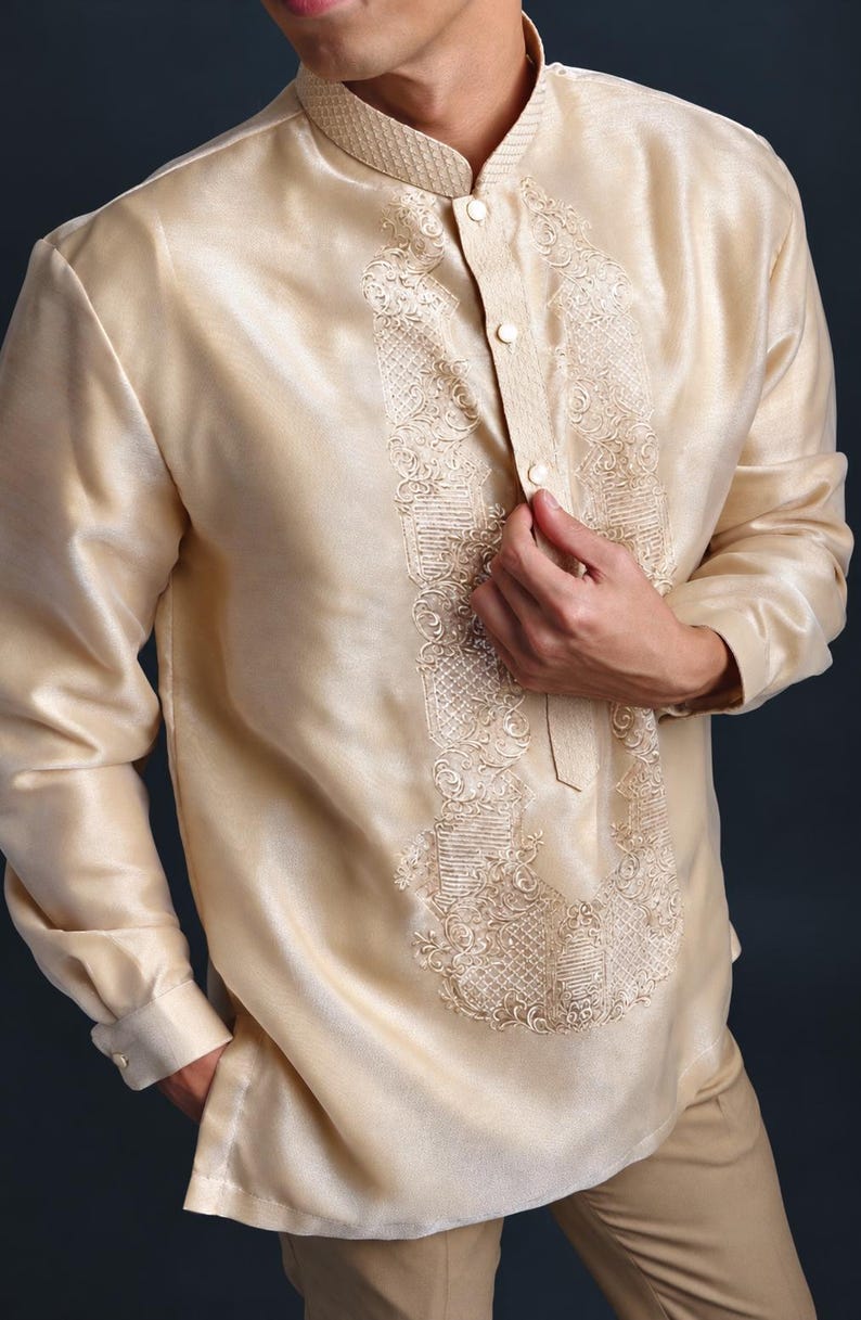 May include: A long-sleeved, champagne-colored barong tagalog shirt featuring a mandarin collar and a central vertical panel with intricate, tonal embroidery. The garment has a button-down front placket and cuffed sleeves, crafted from a semi-sheer, lustrous fabric.