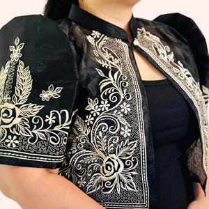May include: Black jacket with intricate white floral embroidery. The jacket features short, puffed sleeves and a mandarin collar. The embroidery includes roses, leaves, and other floral designs, creating a detailed and elegant look.