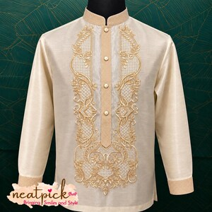 May include: A cream-colored long-sleeve barong shirt featuring a mandarin collar, a button-down placket, and intricate gold embroidery along the front panels. The fabric has a subtle sheen, and the text neatpick shop Bringing Smiles and Style is visible.