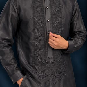 May include: A charcoal gray long-sleeved shirt featuring a mandarin collar, a button-down front placket, and intricate tonal embroidery along the chest panel. The garment is crafted from a semi-sheer fabric with a subtle sheen.