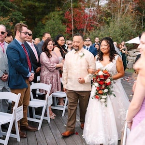 May include: A wedding scene featuring a person in a white lace gown holding a cascading bouquet of red, orange, and white flowers, walking alongside a person in a cream-colored barong tagalog and brown trousers on a wooden deck.