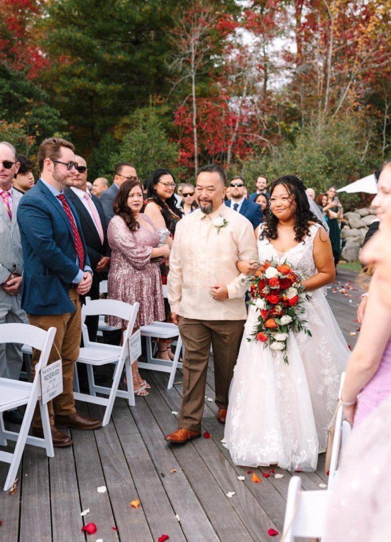 May include: A wedding scene featuring a person in a white lace gown holding a cascading bouquet of red, orange, and white flowers, walking alongside a person in a cream-colored barong tagalog and brown trousers on a wooden deck.