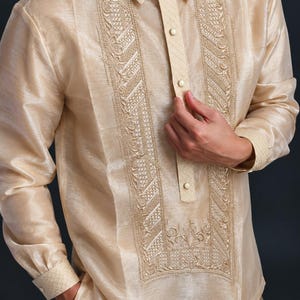 May include: A long-sleeved, champagne-colored barong tagalog shirt featuring a structured collar, a partial button-down placket, and intricate, symmetrical embroidery panels running down the front chest, complemented by matching cuffs.