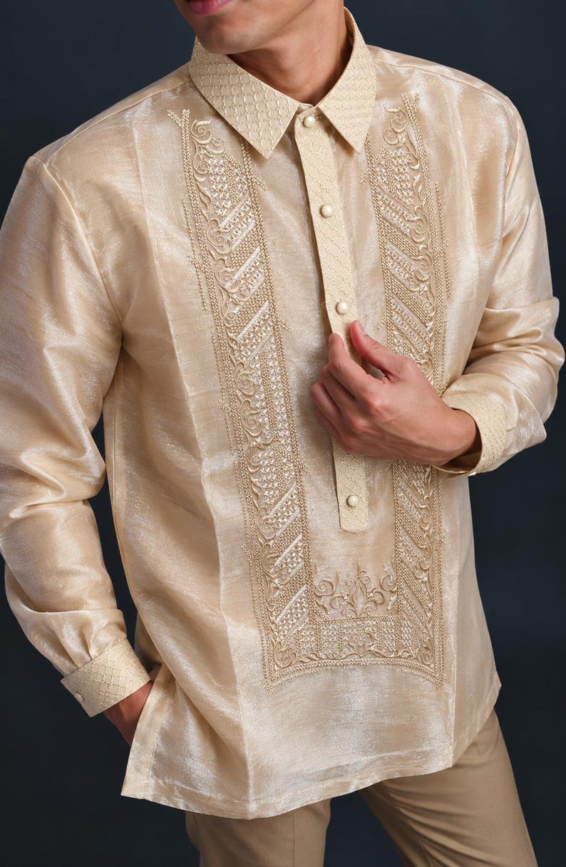 May include: A long-sleeved, champagne-colored barong tagalog shirt featuring a structured collar, a partial button-down placket, and intricate, symmetrical embroidery panels running down the front chest, complemented by matching cuffs.