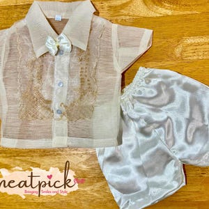 May include: A sheer, cream-colored short-sleeved button-down shirt featuring intricate gold embroidery on the front panels and a small cream bow at the collar, paired with solid white satin shorts that have an elasticized waistband.