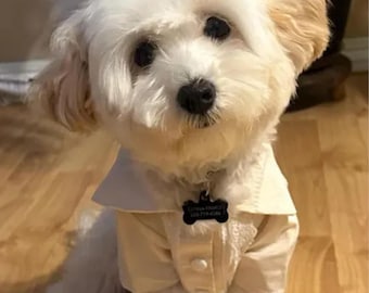 May include: A small white dog with fluffy fur wearing a cream-colored button-down shirt with a collar. The dog has a black bone-shaped identification tag on its collar. The image includes the text neatpick Shop Bringing Smiles and Style.