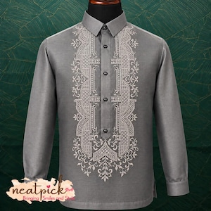 May include: A long-sleeved grey button-down shirt featuring intricate white embroidery along the front placket. The design includes geometric patterns and floral motifs. The shirt has a standard collar and a straight hemline, with a logo in the corner.