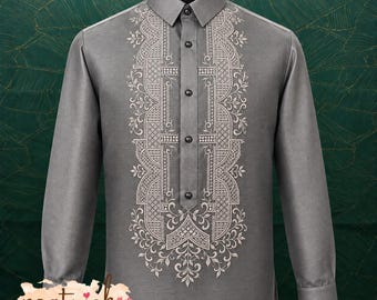 May include: A long-sleeved grey button-down shirt featuring intricate white embroidery along the front placket. The design includes geometric patterns and floral motifs. The shirt has a standard collar and a straight hemline, with a logo in the corner.